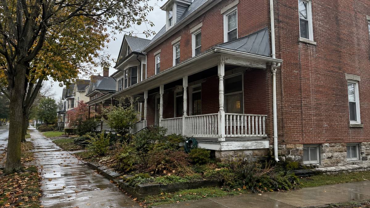 A brick two and a half storey home on a Gettysburg borough street on an overcast autumn afternoon, with a wet sidewalk, fallen leaves and a stone foundation course at ground level.
